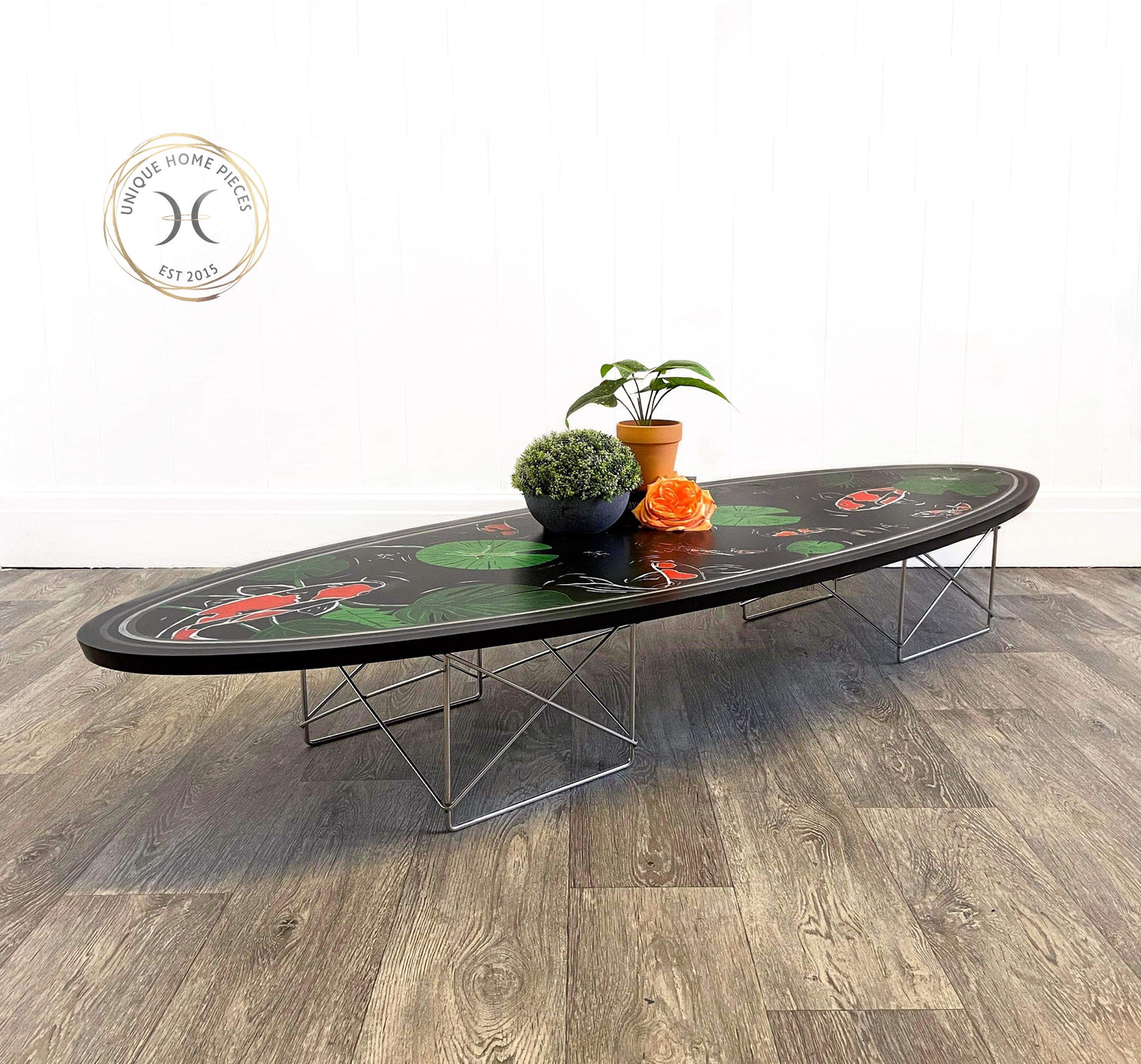 Eames Surfboard Low coffee table with metal leg design low to the floor. Black with a hand painted design applied to the top featuring koi carp in different sizes outlines in with with orange colouring, green pond green flora with insects and a dragonflies..Black grey and silver stripes around the edges to tie in with the chrome base .