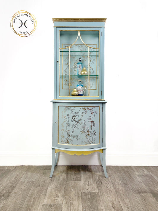 Antique display cabinet consisting of a glazed on three sides cabinet at the top with two new glass shelves at the bottom a good size cupboard with a fixed shelf. All hidden behind a curved door. The cabinet is supported on 4 slightly turned out legs. Painted in pale blue with gold highlighting. A gold transfer depicting birds in trees has been applied to the cabinet back and the cupboard door insert. 